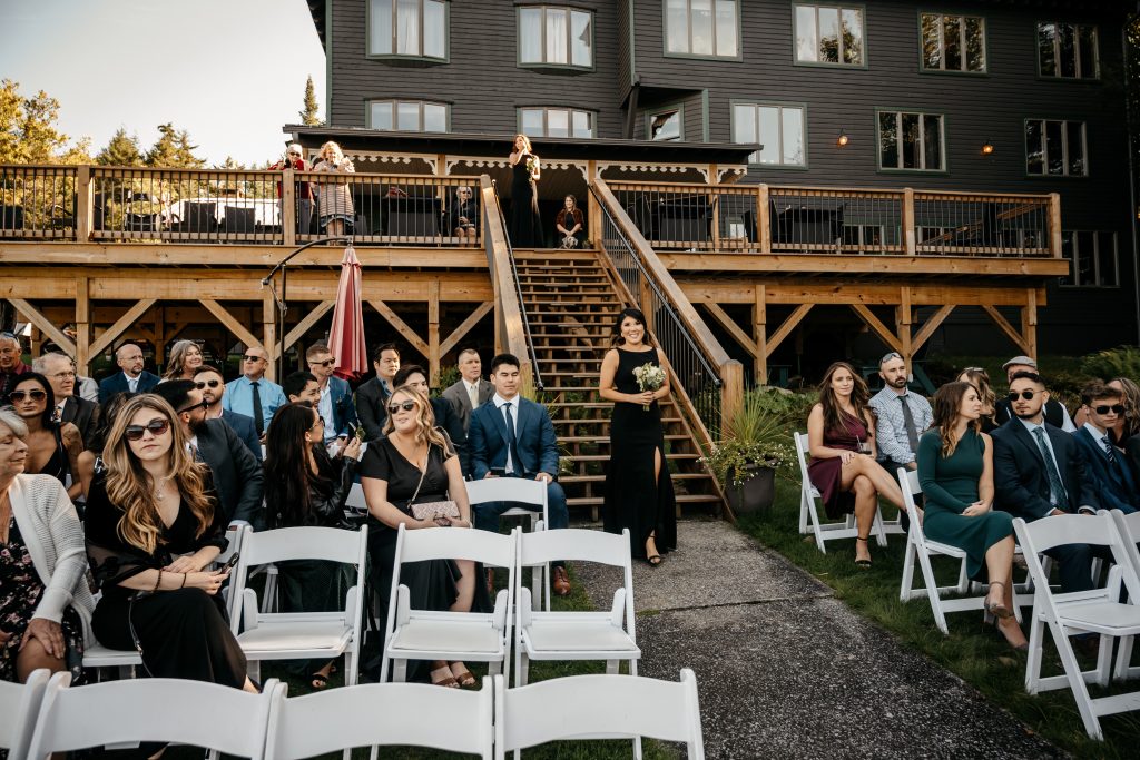 A bridesmaid in a black wedding dress walks down wooden staircase to the alter while a group of guests who don't know the average cost of an upstate ny wedding watch