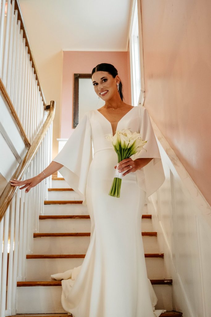 A smiling bride in her wedding dress holds white flowers while standing on a wooden staircase at one of the best Upstate New York wedding venues