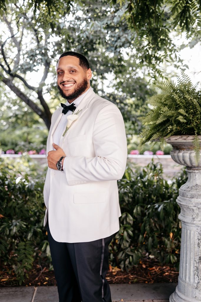 A groom in a white tuxedo stands in a garden and smiles while asking when can you get married in new york