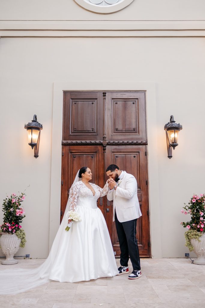 A groom kisse his brides hand in front of tall, wooden doors and flower arrangements while getting married in upstate new york
