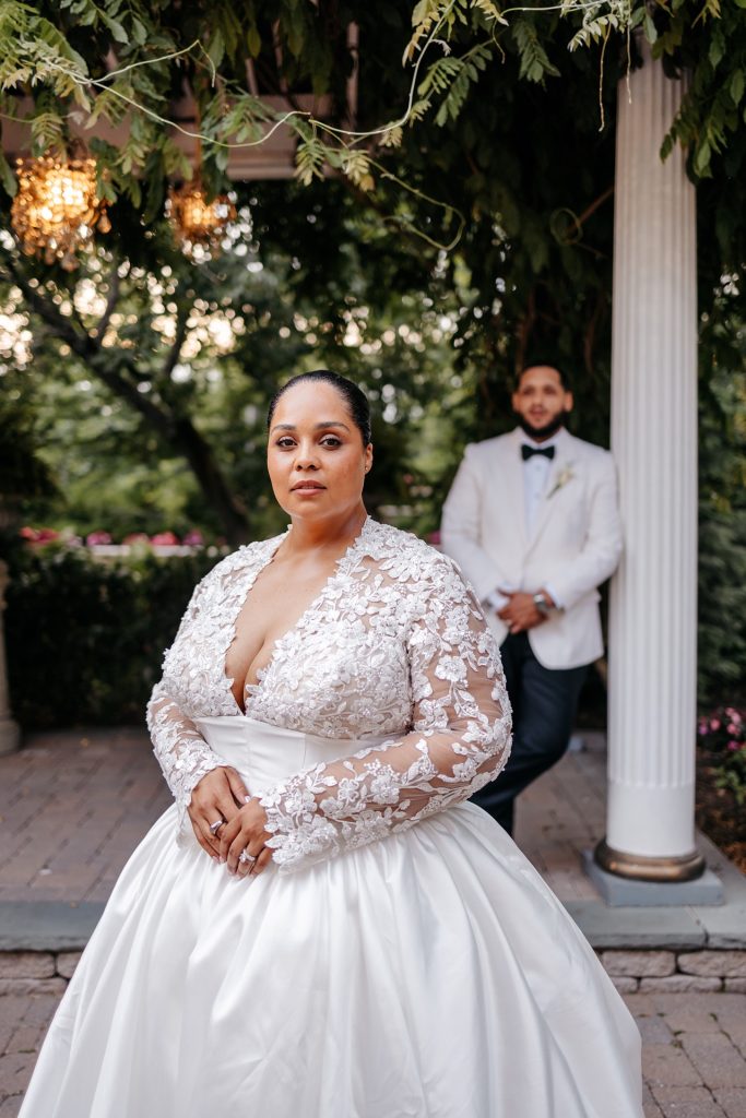 A bride in a lace wedding dress poses for photos with her groom standing in the background wondering what is the best month to get married in Upstate NY