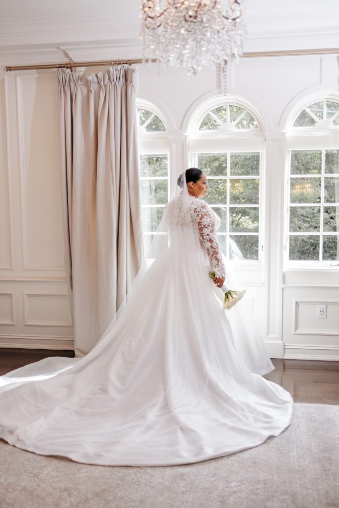 A bride with a long white veil stands in front of a bright window and under a crystal chandelier for a new york wedding photo
