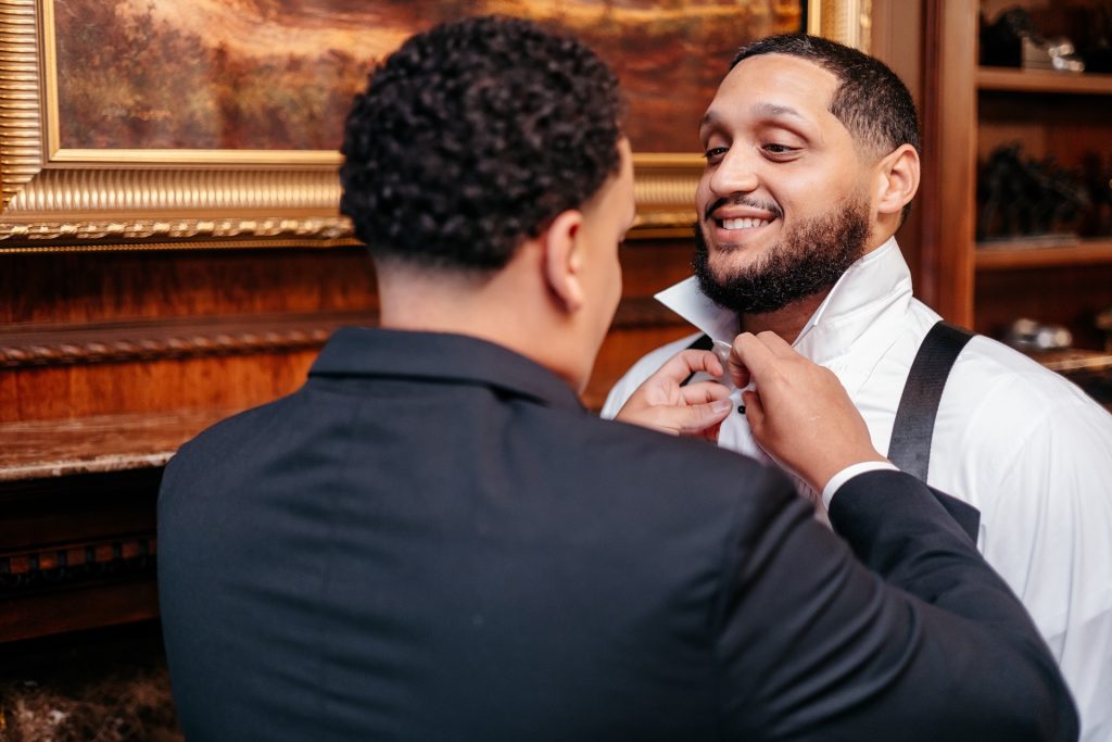 A groom smiles as his best man helps him with his tie for natural wedding photos